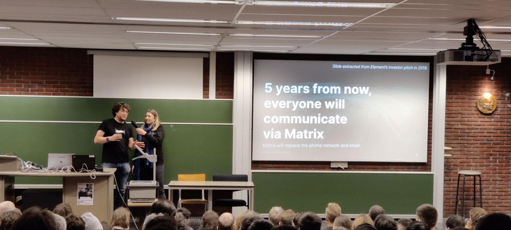 A picture of Matthew & Amandine presenting their slides. Amandine holds the microphone from Matthew. They're in front of slides spelling out "5 years from now, everyone will communicate via Matrix"